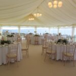 Carpeted wedding marquee with panoramic window walls, globe chandelier lighting and tables set for dining