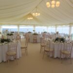 Carpeted wedding marquee with clear panoramic walls, chandelier lighting and set guest tables