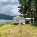 White marquee set up on grass beside a loch. To the left of the marquee is water and hills across the water and to the right are some trees. Another marquee and a white building are visible behind the trees.