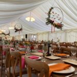 Internal wedding marquee view of lights and circular flower decorations hanging from ceiling above long wooden dining tables set with dark orange and dark pink napkins, plates, cutlery, wine glasses and bottles of wine
