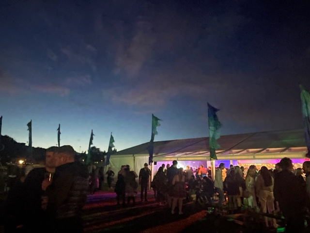 Sundown at the Gig at the Gathering marquee aglow with soft coloured lighting at Cowal Games with crowds, banners and hills