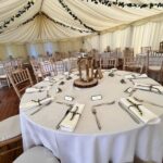 Table settings inside a wedding marquee. Tables are dressed with white cloth covers and napkins and each table has a wooden candle holder with a white candle inside. Chair seat covers are white and the marquee ceiling is decorated with small faux leaves.