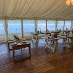 Vintage wooden trestle tables laid with white crockery, glasses and flower pots in marquee with gold balloon-shaped-lights decorating the ceiling. A view of the beach, loch and hills across the water is visible from the windows running the length of the marquee.