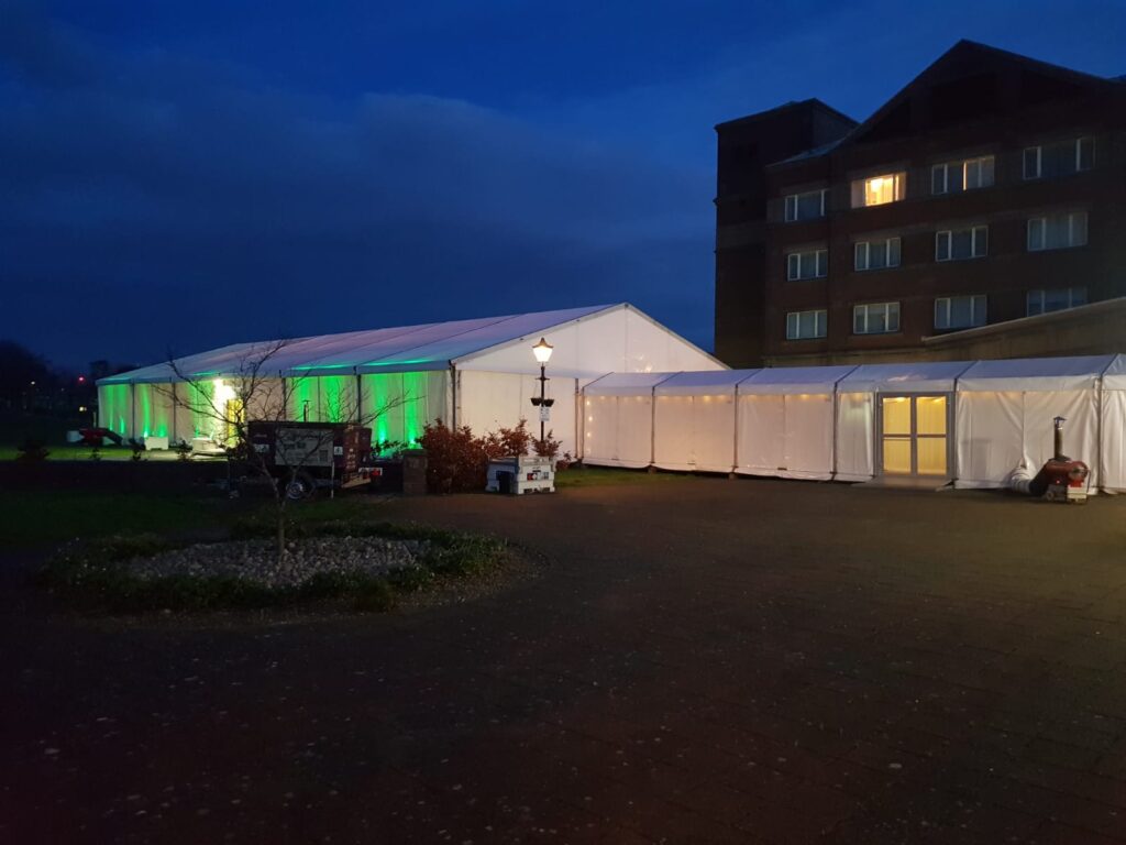 External view of a corporate evening event marquee with LED uplighters along with a marquee link complete with heating and a door.